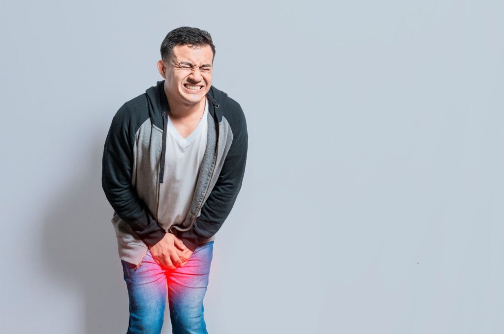 Homem com jeans e moletom segura a região genital com dor, ilustrando lesão peniana aguda com destaque para fratura de pênis.