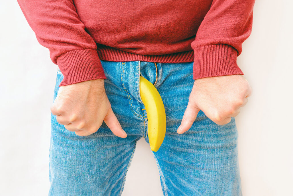 Pessoa de blusa vermelha e jeans aponta para um elemento amarelo curvo que representa a curvatura peniana.