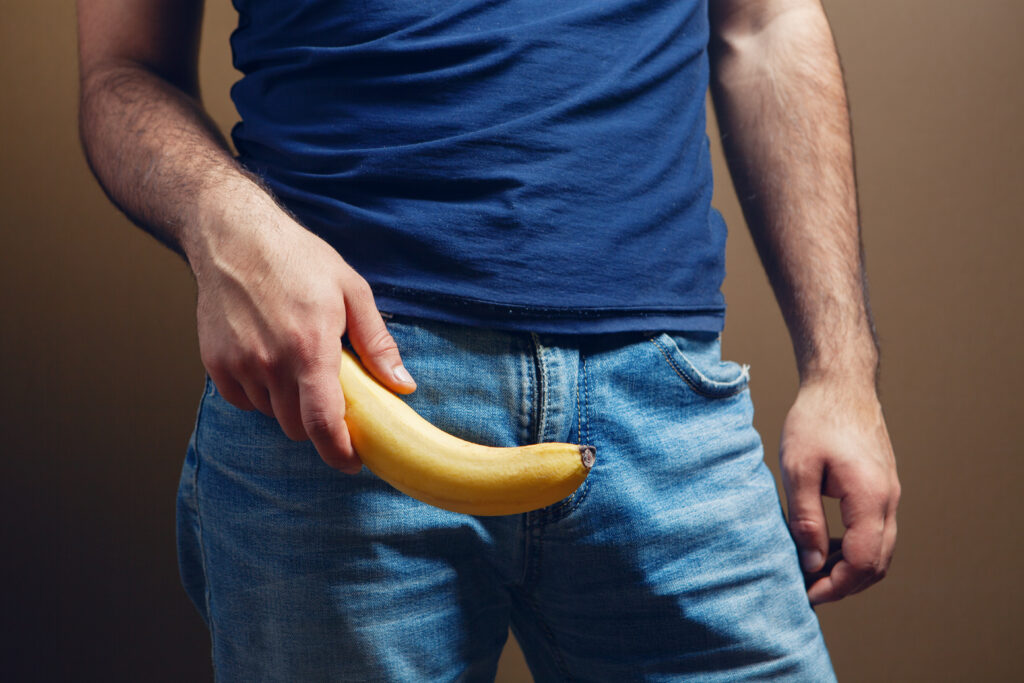 Homem segurando uma banana sobre a região genital, em uma representação visual de um pênis curvo para ilustrar a Doença de Peyronie e a curvatura peniana.