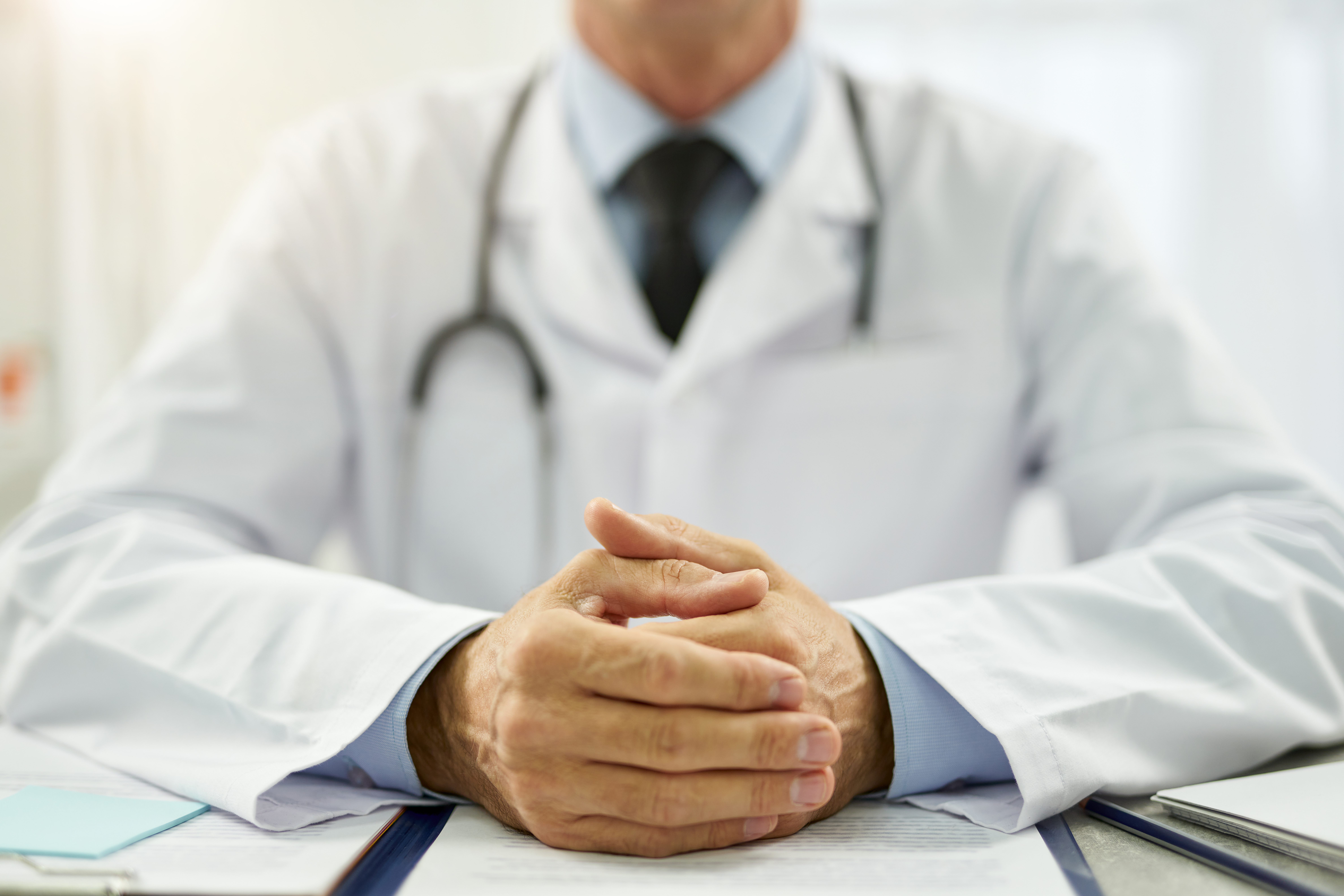 Médico de jaleco branco e estetoscópio mantém as mãos entrelaçadas sobre uma mesa com documentos, em ambiente clínico claro.