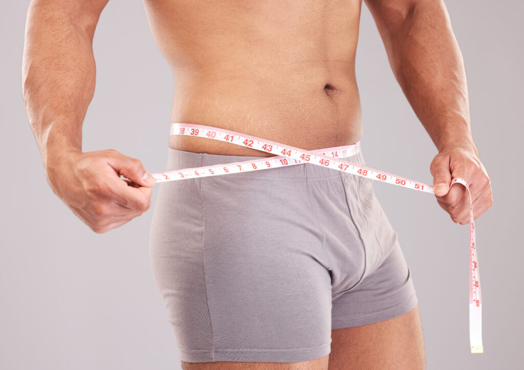 Homem sem camisa e de roupa íntima cinza mede a circunferência da cintura com uma fita métrica branca, segurando as duas extremidades com as mãos.