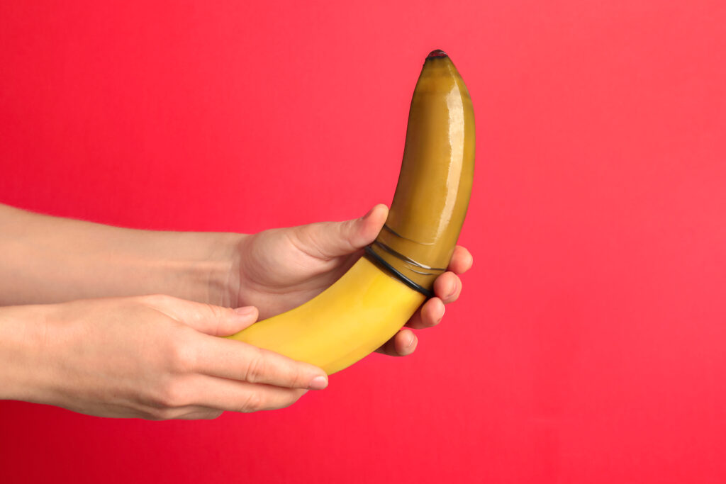 Mãos segurando uma banana amarela com um preservativo desenrolado, representando visualmente o tema do pênis curvo e da doença de Peyronie.