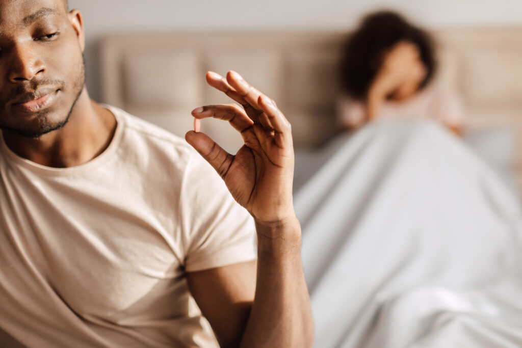 Homem sentado na cama segura um comprimido entre os dedos enquanto observa o medicamento, com uma pessoa desfocada ao fundo, ilustrando o tratamento da disfunção erétil e a importância da avaliação médica.