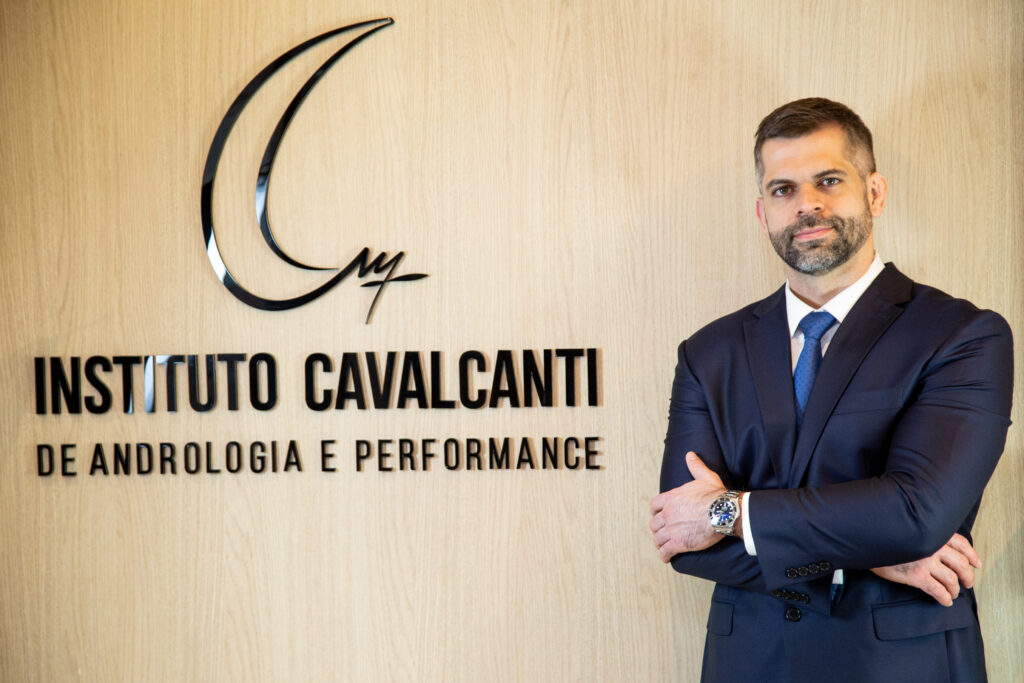 Homem de terno azul posa de braços cruzados diante da parede com o logotipo e o nome “Instituto Cavalcanti de Andrologia e Performance”.
