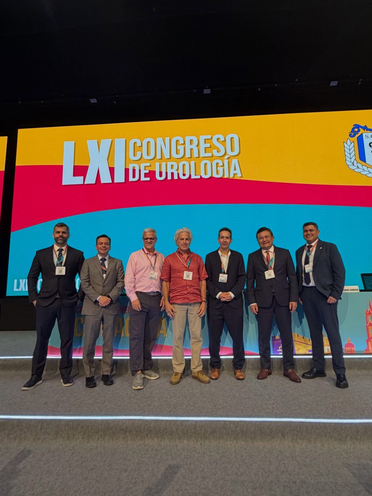 Sete participantes posam no palco do LXI Congresso de Urologia diante de um grande painel colorido do evento; Dr. Marco Túlio aparece na extremidade esquerda do grupo.