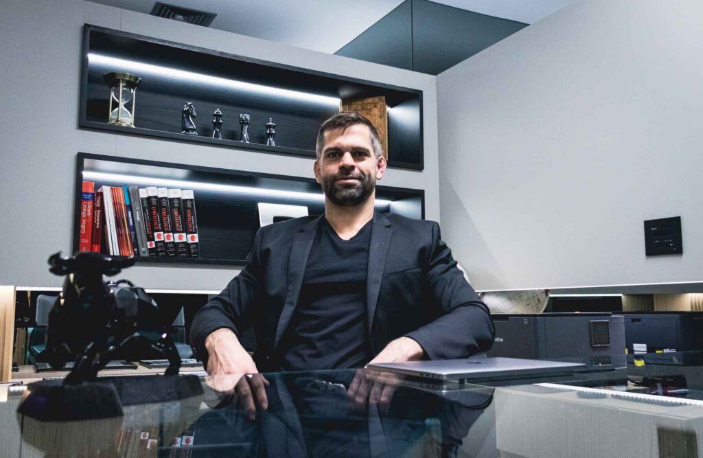 Dr. Marco Túlio Cavalcanti, andrologista no Itaim Bibi, em São Paulo, posa sentado à mesa de seu consultório, vestindo blazer preto e camiseta escura. Ao fundo, aparecem prateleiras iluminadas com livros de referência médica, objetos decorativos e equipamentos de escritório. A fotografia destaca o médico em seu ambiente profissional e está relacionada à atuação em saúde masculina, diagnóstico de condições urológicas e andrológicas, avaliação clínica e tratamentos individualizados.